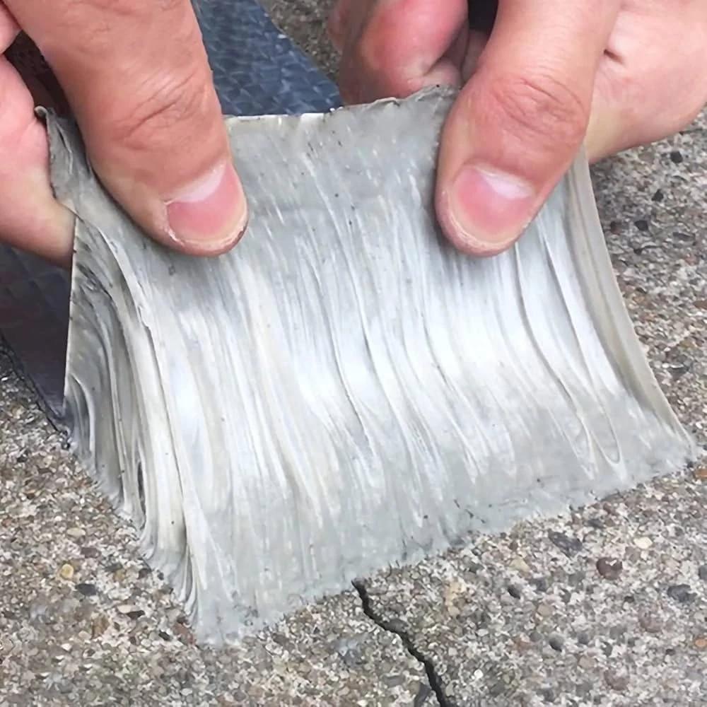 Two hands peeling the tape back from a concrete path, the grey butyl underside stretching into long strings that show how thick and tacky the sealant layer is.